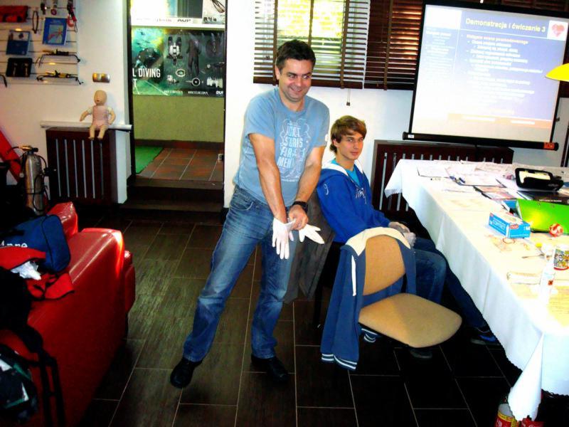 Instruktor w Przeźmierowie demonstruje zakładanie rękawic ochronnych podczas szkolenia z pierwszej pomocy przedmedycznej; w tle siedzący uczestnik i ekran projekcyjny z prezentacją.