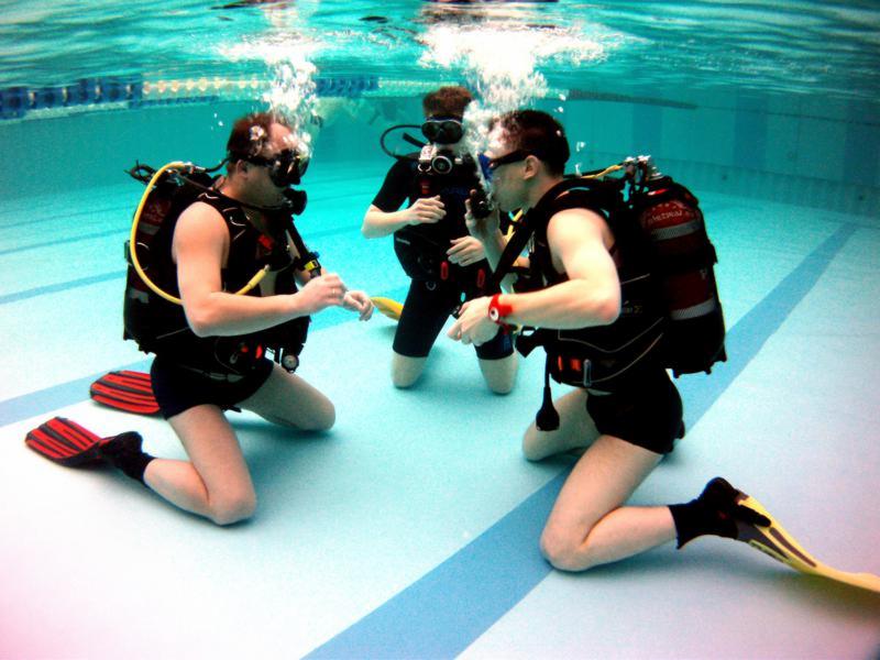 Trzech mężczyzn w strojach do nurkowania klęczy na dnie basenu, wypuszczając powietrze z automatów oddechowych, widoczne bąbelki powietrza, czerwone i żółte płetwy.