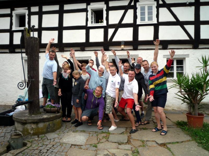 Grupa roześmianych osób pozuje z podniesionymi rękami na tle budynku z muru pruskiego i starej studni z kwiatami.