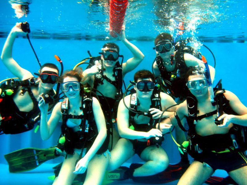 Grupa osób w maskach i sprzęcie do nurkowania, znajdująca się pod wodą w basenie, z widocznymi bąbelkami powietrza i czerwoną liną oddzielającą tory.