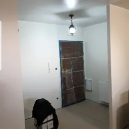 Wnętrze w trakcie prac: świeżo pomalowane ściany na biało, zabezpieczone drzwi folią, podłoga z taśmą malarską, lampa sufitowa w stylu retro, pudełka farby przy ścianie.