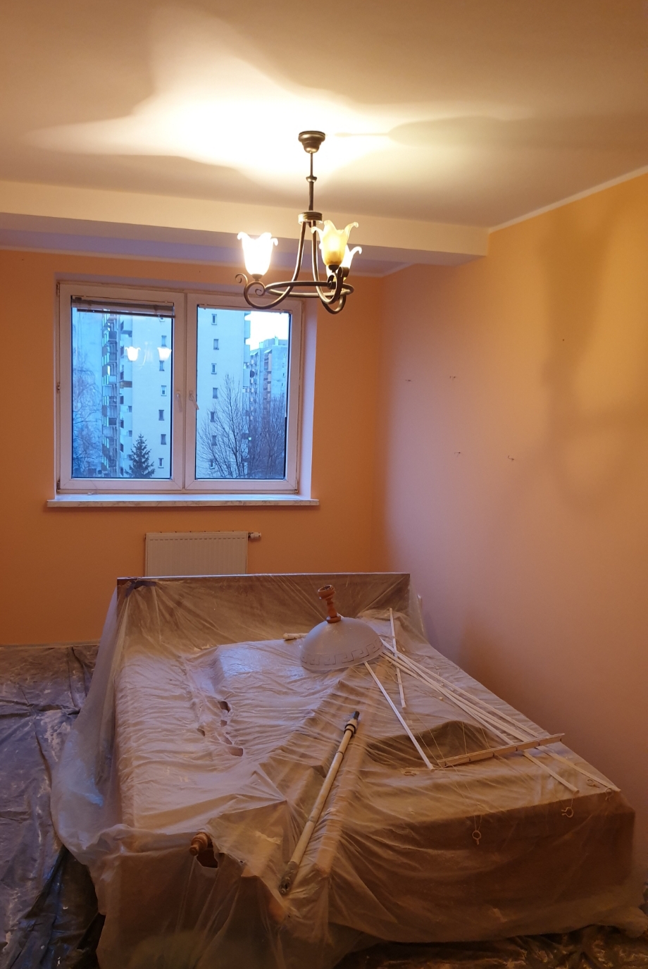Pokój w trakcie zabezpieczania przed pracami wykończeniowymi, meble przykryte folią ochronną, widoczne ślady po mocowaniach na pomarańczowej ścianie, okno z widokiem na blokowisko, lampa wisząca.