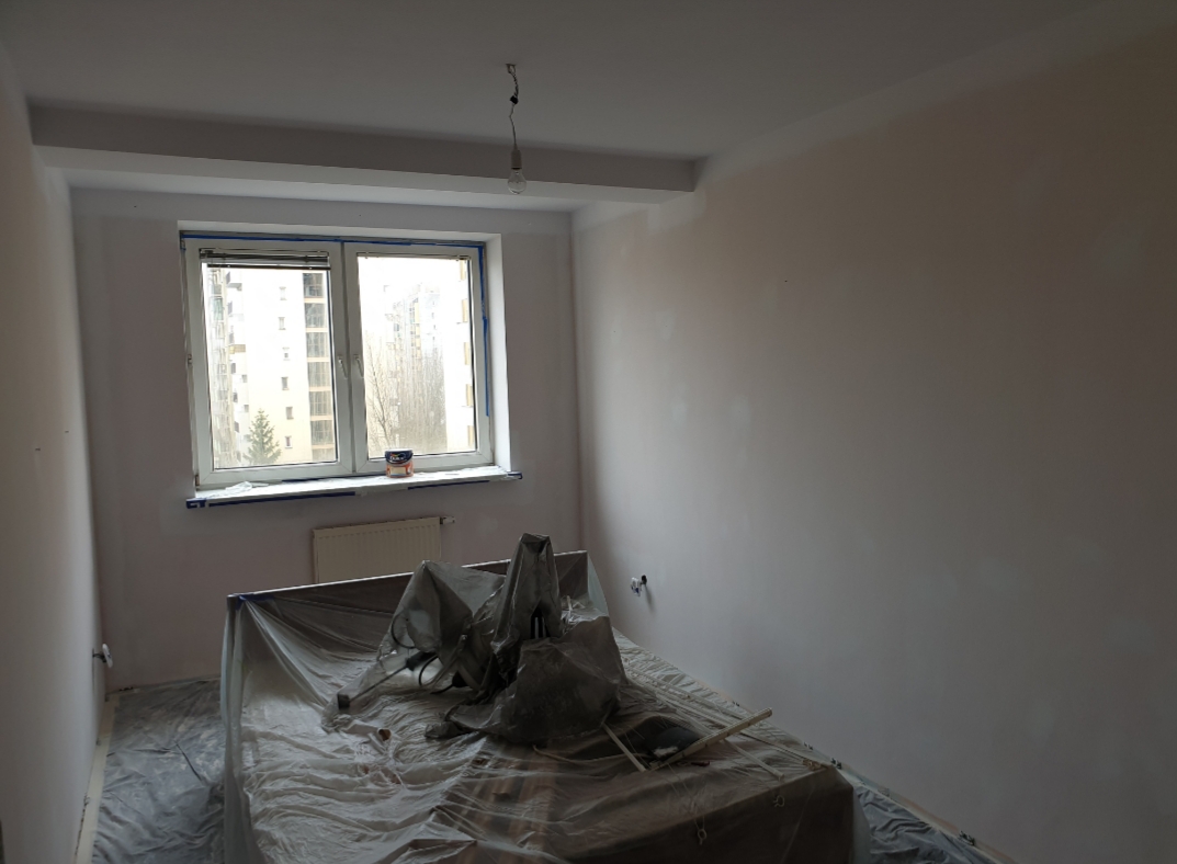 Pokój w trakcie odświeżania, ściany pomalowane na jasny kolor, meble zabezpieczone folią malarską, widoczne ślady po szpachlowaniu na ścianach, okno z widokiem na blok.