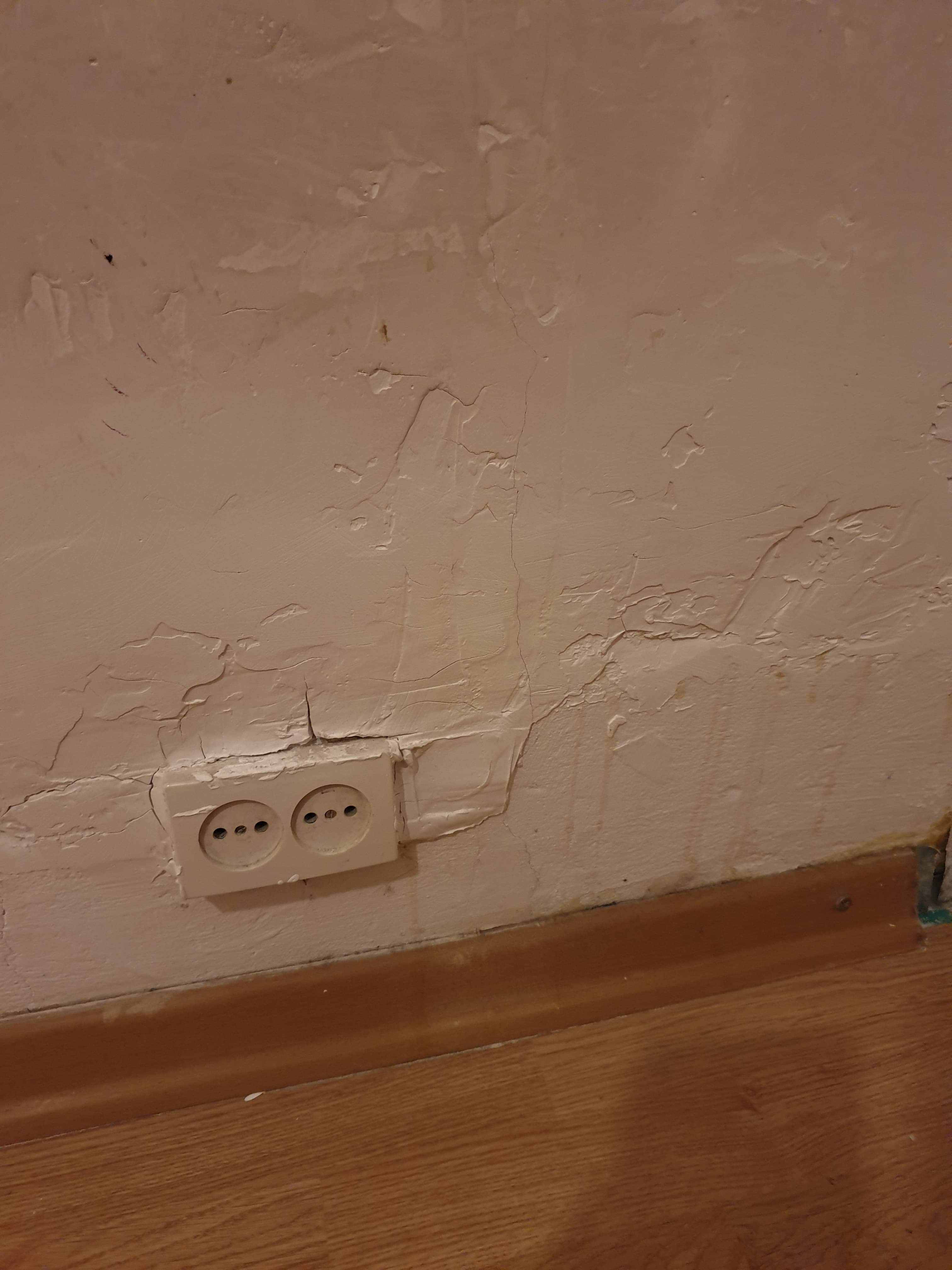 Nierówna, popękana powierzchnia ściany z gniazdkiem elektrycznym, widoczne ślady po naprawach i malowaniu, brudna podłoga z drewnianą listwą przypodłogową.