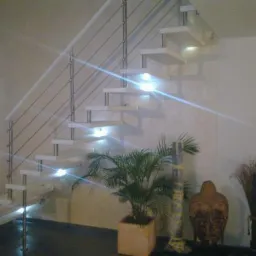 Nowoczesne schody z białymi stopniami i metalową balustradą w jasnym wnętrzu, oświetlone punktowo, z dekoracyjnymi roślinami i rzeźbą u podstawy.