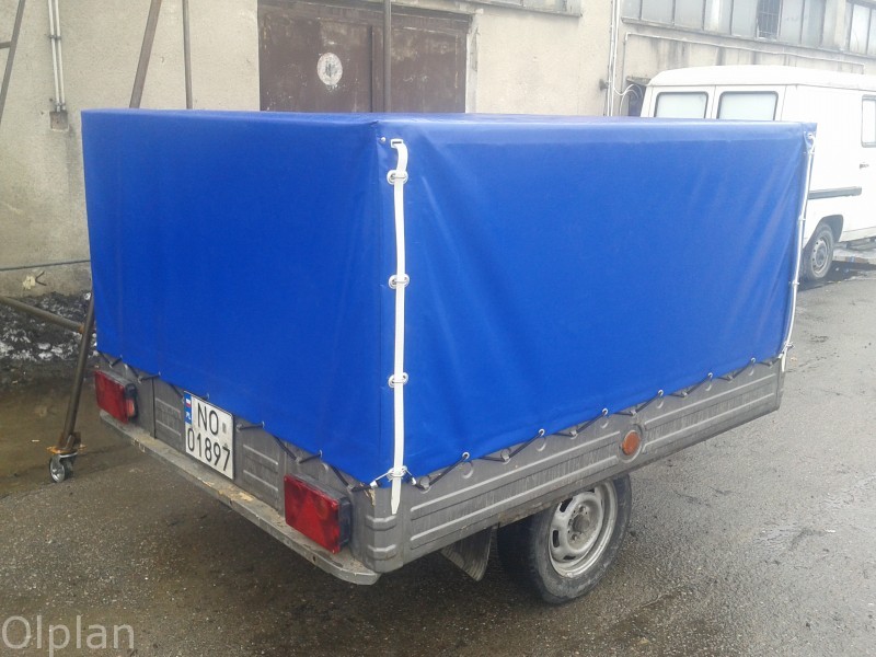 Niebieska plandeka na przyczepce samochodowej, widoczna tablica rejestracyjna i tylne światła, tło stanowi budynek przemysłowy i zaparkowany biały bus.