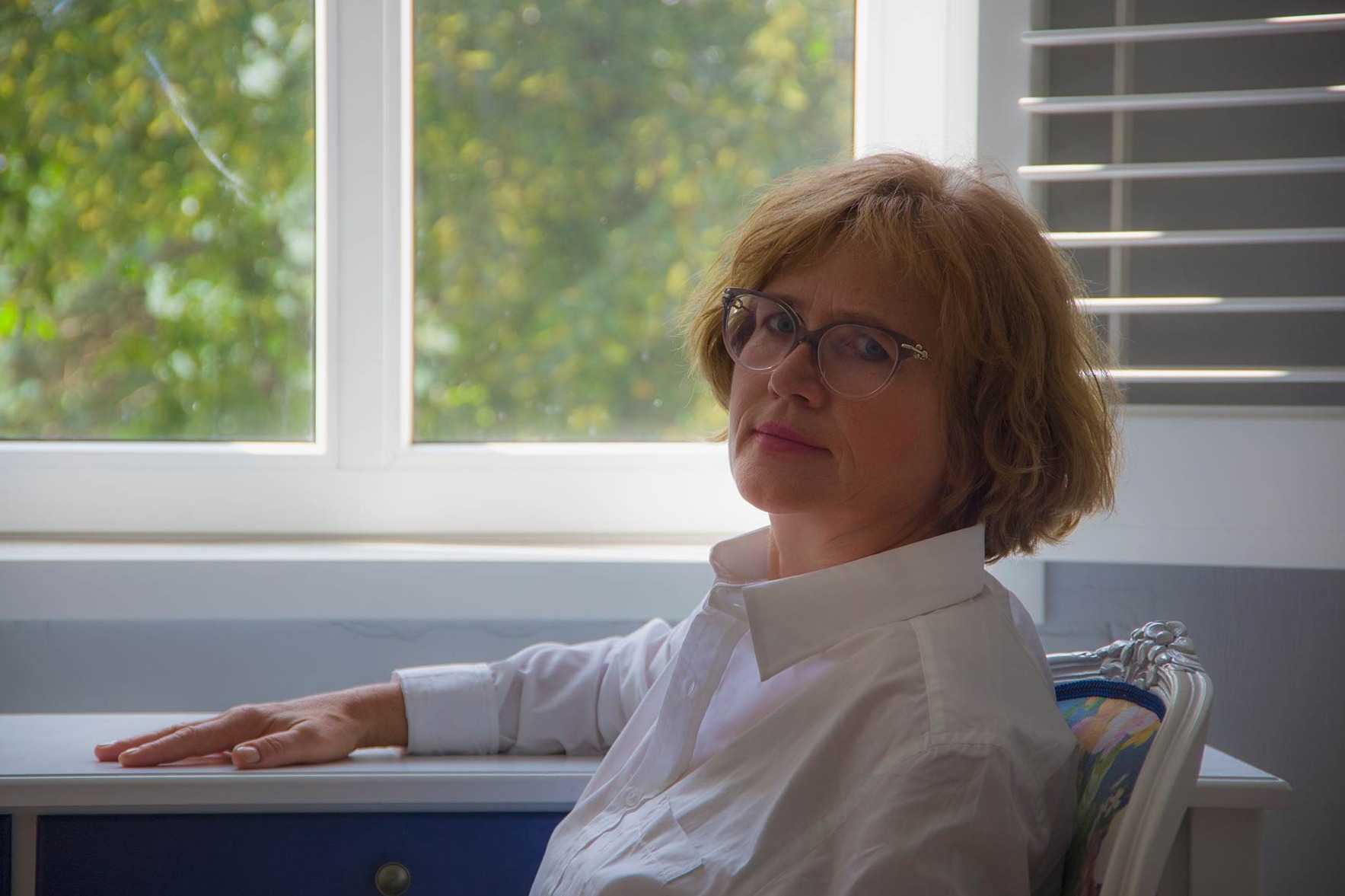Portret kobiety w okularach w gabinecie w Chojnicach. Ubrana w białą koszulę, siedzi przy biurku na ozdobnym krześle, z oknem i żaluzjami w tle.