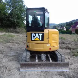 Żółta mini koparka CAT na placu budowy, widok z przodu, w tle domy i zalesione wzgórza.