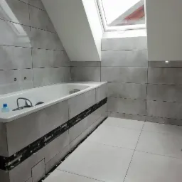 Wykończona łazienka na poddaszu z szarymi, poziomymi płytkami, białą wanną zabudowaną płytkami z dekoracyjnym pasem, oraz oknem dachowym.