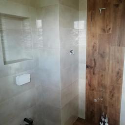 Dariusz Górka - Wykończona łazienka w Żołyni z beżowymi płytkami imitującymi kamień, dekoracyjną wnęką i fragmentem ściany z płytek drewnopodobnych, widoczne akcesoria montażowe.