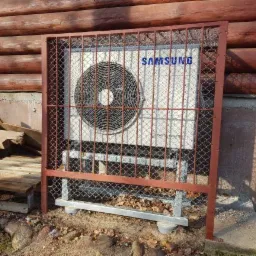 Zewnętrzna jednostka klimatyzacji Samsung, zabezpieczona metalową kratą, umieszczona na betonowej podstawie przy drewnianym budynku.
