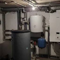 Instalacja grzewcza w piwnicy, widoczne zbiorniki, rury, zawory i urządzenia sterujące, ściany z surowym wykończeniem.