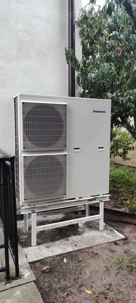 Zewnętrzna jednostka klimatyzacyjna Panasonic na białej, metalowej konstrukcji, umieszczona przy szarej ścianie budynku z widocznym brązowym rynną i zielonym drzewem w tle.