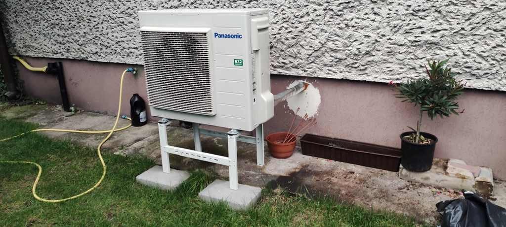 Zewnętrzna jednostka pompy ciepła Panasonic zamontowana na białym stelażu na betonowych podstawach, z widocznymi podłączeniami hydraulicznymi i butelką płynu obok, na tle ściany budynku z fakturą...