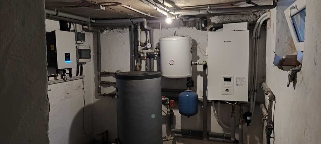 Instalacja grzewcza w piwnicy, widoczne zbiorniki, rury, zawory i urządzenia sterujące, ściany z surowym wykończeniem.
