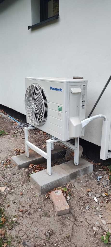 Zewnętrzna jednostka klimatyzacji Panasonic R32 zamontowana na białym metalowym stelażu na betonowych podstawach przy białej ścianie budynku.