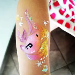 Dziecięce ramię z namalowanym brokatowym jednorożcem w odcieniach różu, fioletu i złota, otoczonym białymi gwiazdkami.