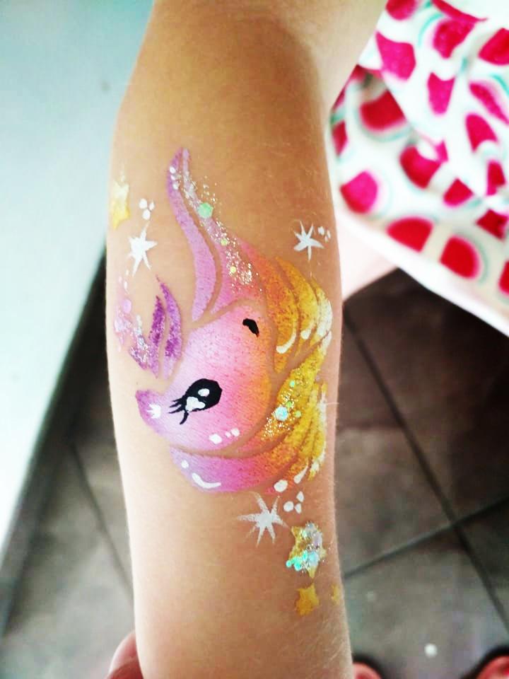 Dziecięce ramię z namalowanym brokatowym jednorożcem w odcieniach różu, fioletu i złota, otoczonym białymi gwiazdkami.