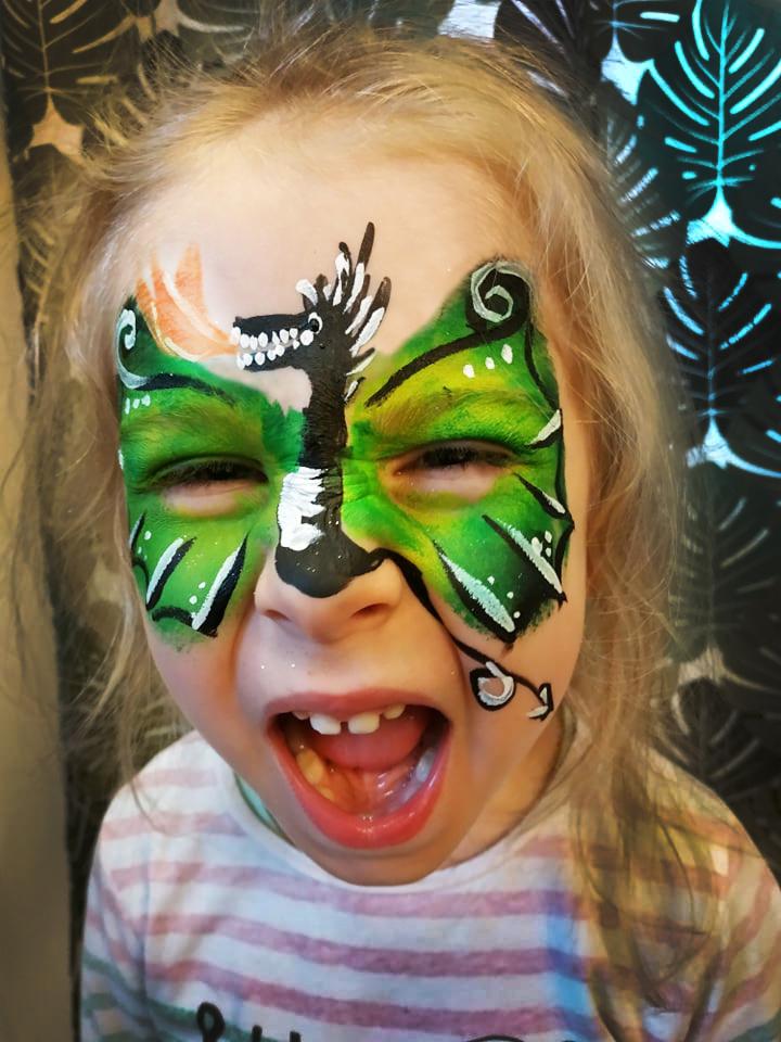 Dziewczynka z pomalowaną twarzą w motyw smoka z zielonymi skrzydłami i czarnym ciałem, z otwartymi ustami i widocznymi zębami, na tle wzorzystej zasłony.