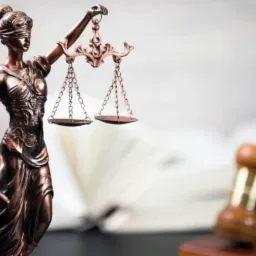 Brązowa statuetka Temidy z wagą i mieczem na tle rozłożonej książki i drewnianego młotka sędziowskiego.