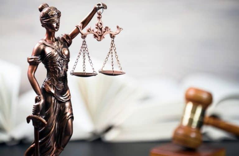 Brązowa statuetka Temidy z wagą i mieczem na tle rozłożonej książki i drewnianego młotka sędziowskiego.