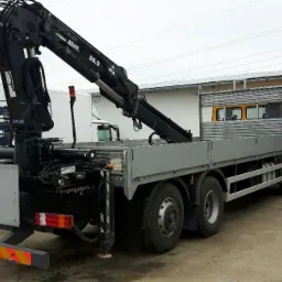 Żółta ciężarówka z zamontowanym czarnym dźwigiem Atlas 25.2 na szarej platformie, zaparkowana na placu z widocznym budynkiem w tle.