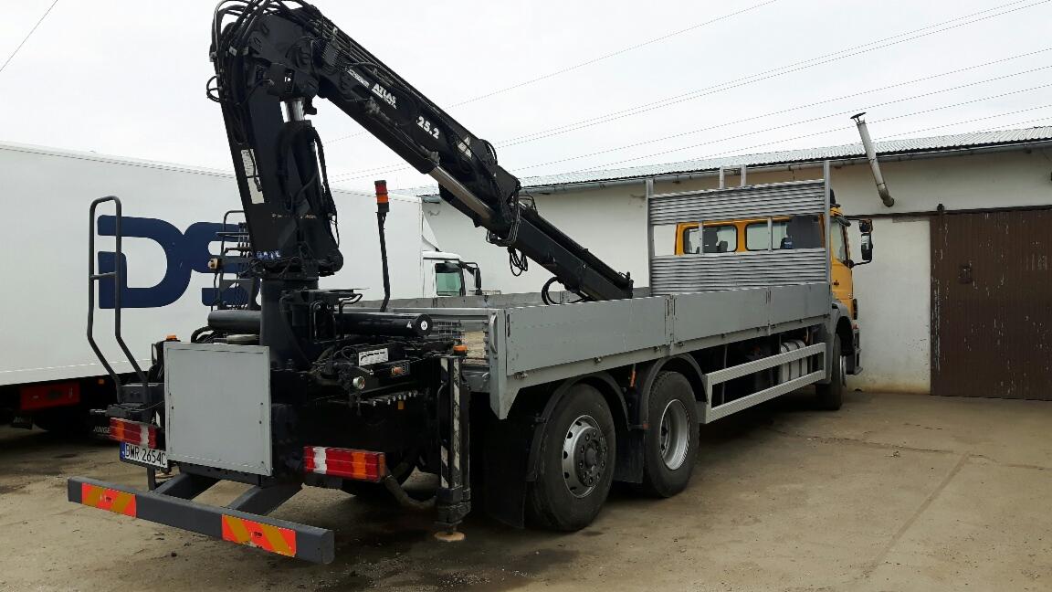 Żółta ciężarówka z zamontowanym czarnym dźwigiem Atlas 25.2 na szarej platformie, zaparkowana na placu z widocznym budynkiem w tle.