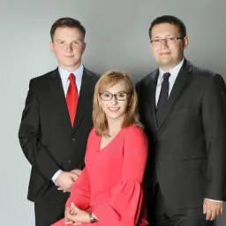 Portret trzech prawników na szarym tle: kobieta w okularach i czerwonej sukience siedzi na pierwszym planie, dwaj mężczyźni w garniturach stoją za nią.