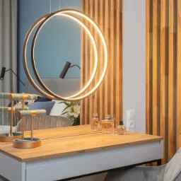 Toaletka z drewnianym blatem, szarą tapicerowaną krzesłem i okrągłym podświetlanym lustrem, na blacie flakoniki perfum i stojak na biżuterię, w tle drewniana ażurowa ścianka działowa.