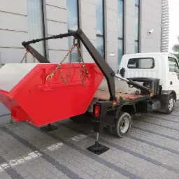 Czerwony kontener na gruz zawieszony na łańcuchach nad platformą białej ciężarówki na tle szarego budynku z dużymi oknami.