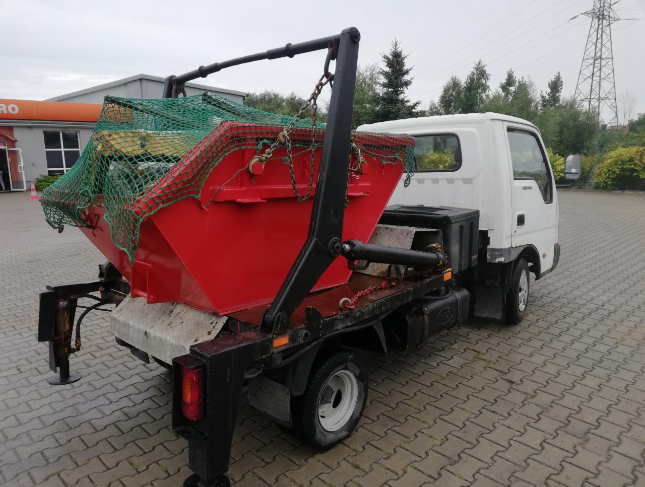 Czerwony kontener na gruz, zabezpieczony zieloną siatką, umieszczony na platformie białego samochodu ciężarowego z wysięgnikiem, na tle parkingu z kostki brukowej i budynków.