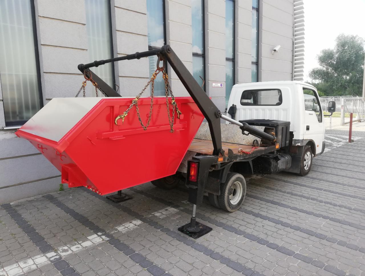 Czerwony kontener na gruz zawieszony na łańcuchach nad platformą białej ciężarówki na tle szarego budynku z dużymi oknami.