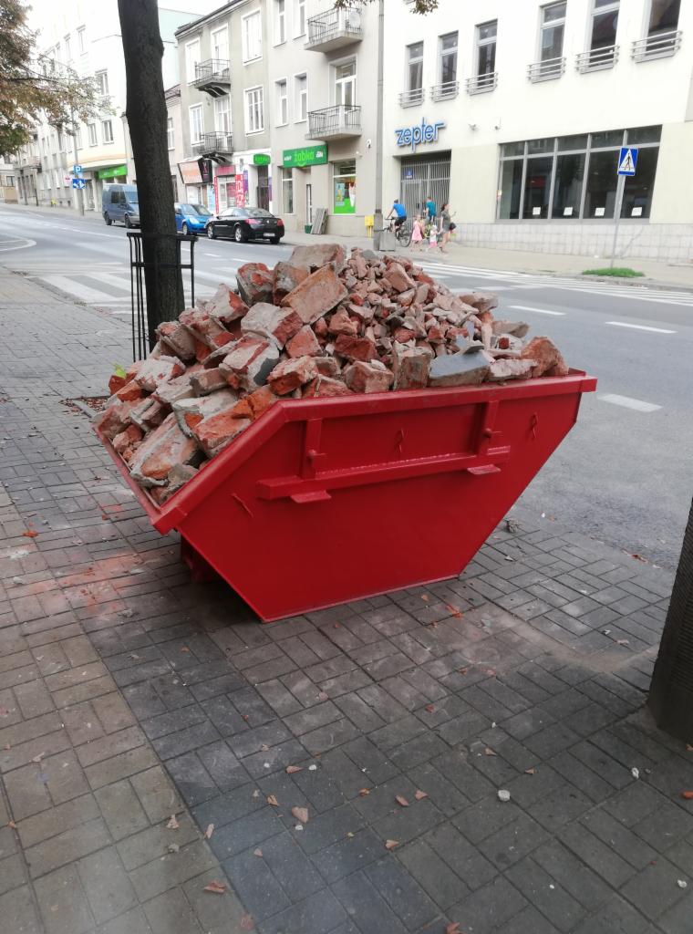 Czerwony kontener wypełniony gruzem ceglanym stoi na chodniku w miejskiej scenerii, z budynkami i przejściem dla pieszych w tle.