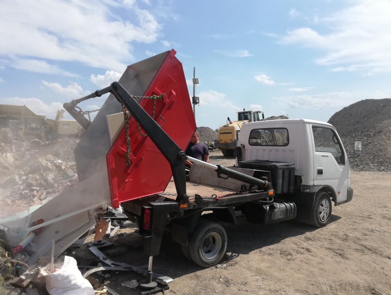 Ciężarówka z czerwonym kontenerem przechyla się, wysypując gruz; w tle koparka i hałda ziemi.