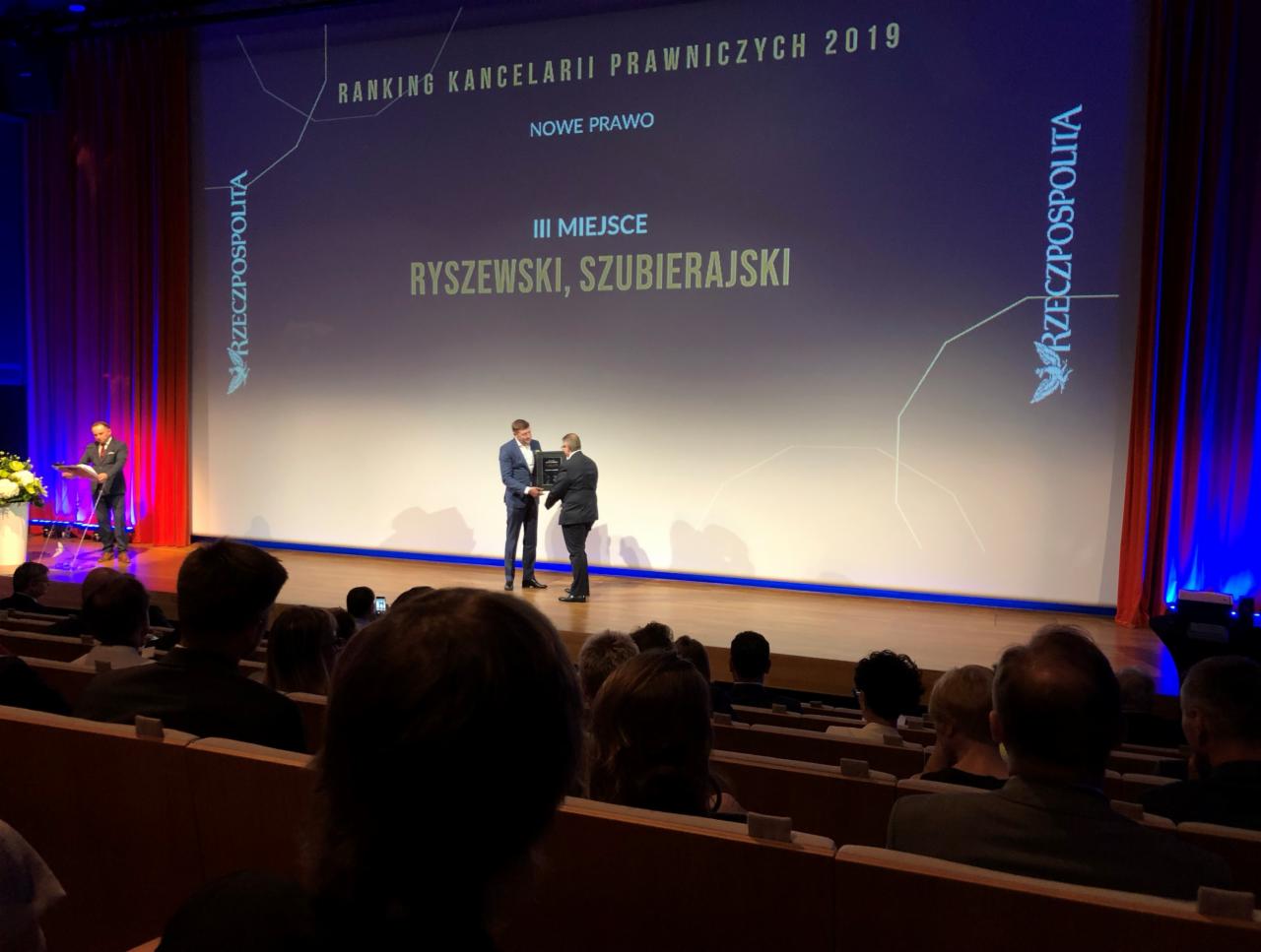 Uroczystość wręczenia nagród w rankingu kancelarii prawnych, widok z publiczności na scenę, gdzie dwaj mężczyźni w garniturach wymieniają się statuetką na tle ekranu z napisem 'Ranking Kancelarii...