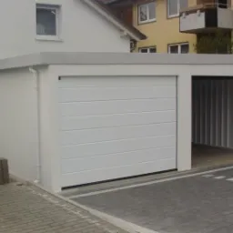 Nowoczesny, dwustanowiskowy garaż z białymi, poziomymi panelami na bramie, z otwartą przestrzenią po prawej stronie, ukazującą metalową konstrukcję wnętrza, na tle jasnych budynków mieszkalnych...