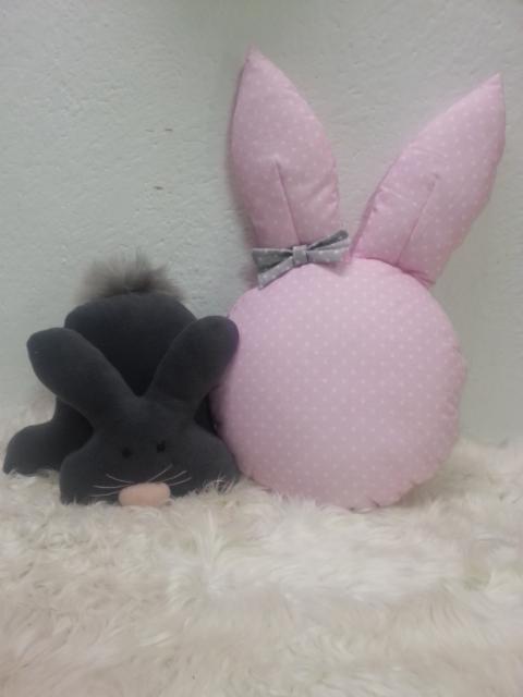 Dwa pluszowe króliki: szary królik siedzący obok różowej poduszki w kształcie głowy królika z uszami w kropki i szarą kokardką, leżące na białym puszystym dywanie.