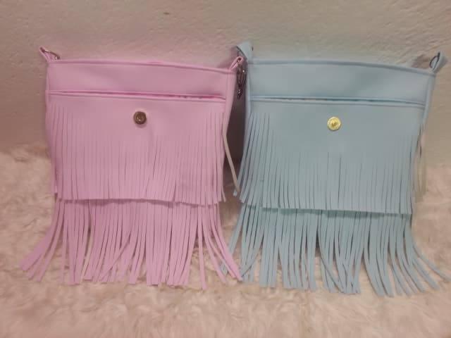 Dwie małe torebki z frędzlami, jedna różowa, druga błękitna, leżą na białym, puszystym materiale.