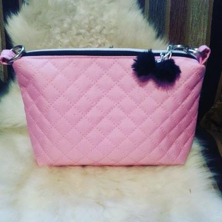 Różowa, pikowana kosmetyczka z czarnymi pomponami przy suwaku, leżąca na puszystym, białym futerku.