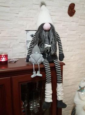 Ręcznie wykonana, dekoracyjna lalka krasnal w czarno-białe paski, siedząca na ciemnobrązowej komodzie. Krasnal ma długą, szarą brodę i trzyma mniejszego krasnala. W tle biała, teksturowana ściana.