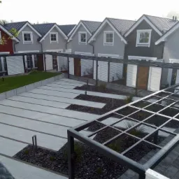 Szereg nowo wybudowanych domów jednorodzinnych z szarymi dachami i różnymi odcieniami elewacji, połączonych metalową konstrukcją zadaszenia nad parkingami, z wykończonym terenem z kostki i czarnego...