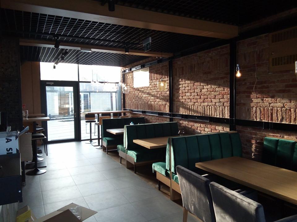Wnętrze lokalu usługowego z ceglaną ścianą, zielonymi lożami i drewnianymi stolikami, widok na zewnątrz przez przeszklone drzwi.