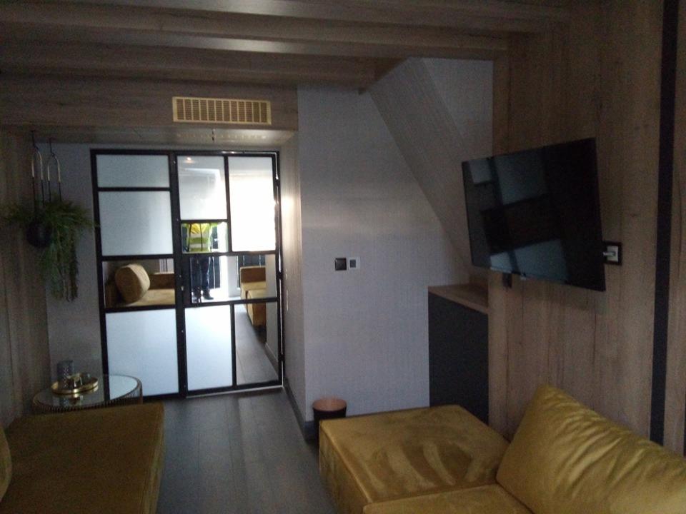 Salon z żółtą sofą i czarnymi, przeszklonymi drzwiami przesuwnymi w stylu loftowym, z widoczną rośliną wiszącą i telewizorem na drewnianej ścianie.