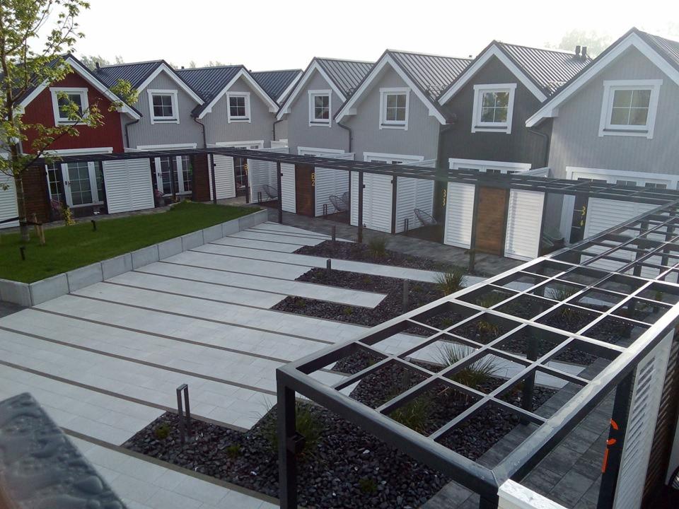Szereg nowo wybudowanych domów jednorodzinnych z szarymi dachami i różnymi odcieniami elewacji, połączonych metalową konstrukcją zadaszenia nad parkingami, z wykończonym terenem z kostki i czarnego...