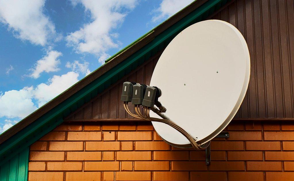 Biała antena satelitarna zamontowana na ceglanej ścianie domu z brązową drewnianą elewacją, pod błękitnym niebem z chmurami.