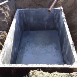 Betonowy zbiornik, prawdopodobnie szambo, umieszczony w wykopie ziemnym, widoczne wystające elementy konstrukcyjne i rura odpływowa.