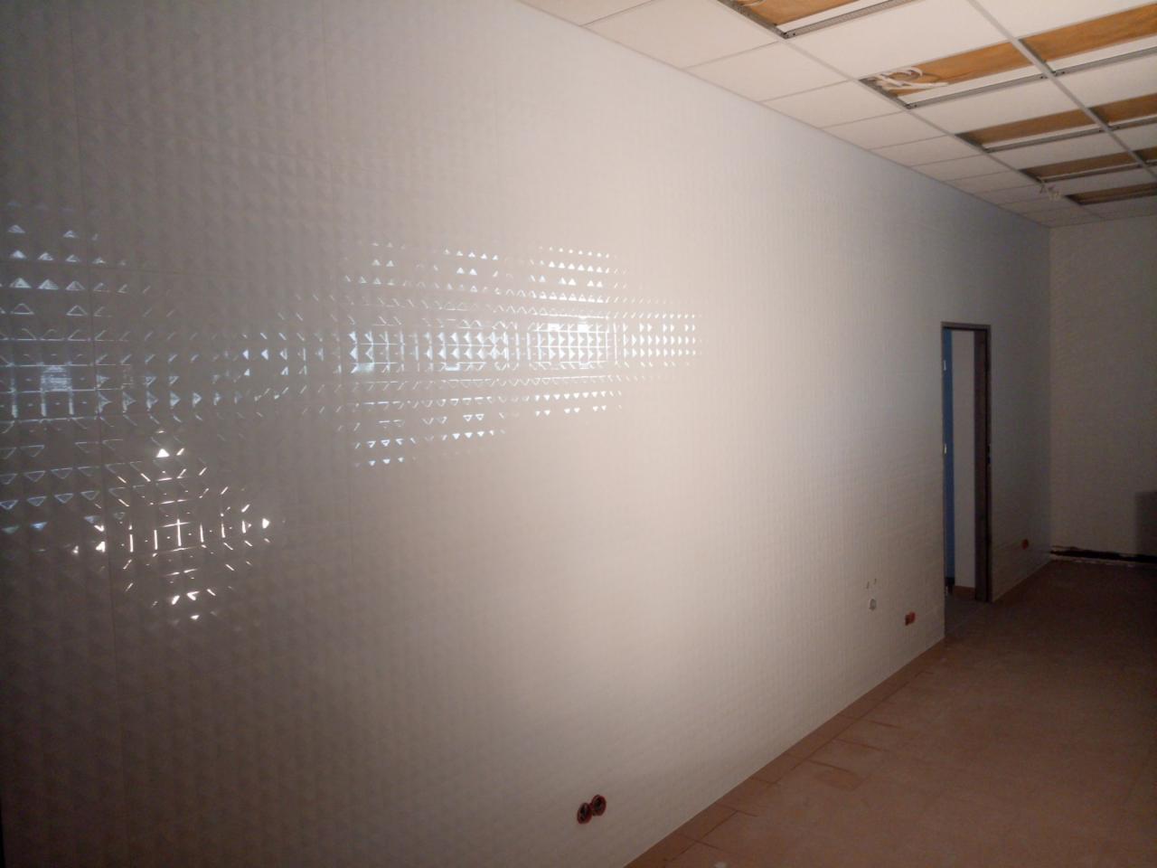 Biała ściana wyłożona błyszczącymi, trójkątnymi płytkami z efektem 3D, widoczne niedokończone prace przy suficie podwieszanym i instalacji elektrycznej.