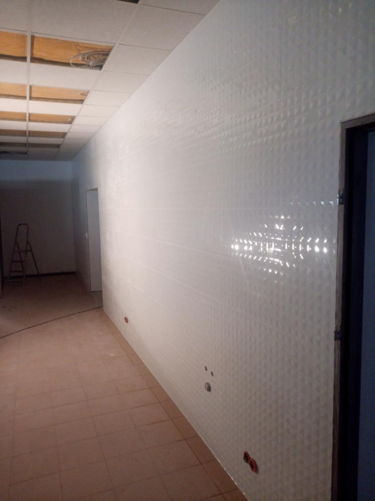 Długa ściana wyłożona białymi, trójwymiarowymi płytkami ceramicznymi w trakcie prac wykończeniowych; widoczne otwory na gniazdka elektryczne, sufit podwieszany z odsłoniętą konstrukcją, drabina w tle.