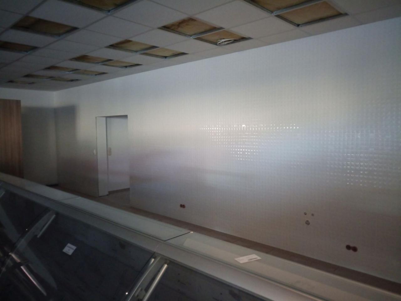 Biała ściana z delikatną fakturą kwadratów w nowym lokalu, widoczne ślady po instalacjach elektrycznych, sufit kasetonowy w trakcie montażu, wnętrze w stanie surowym.