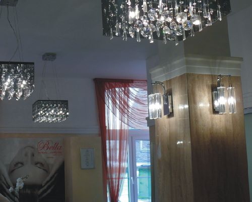 Wnętrze z trzema wiszącymi, kwadratowymi lampami z kryształkami, duży żyrandol z kryształkami, dwa kinkiety na kolumnie i plakat na ścianie.